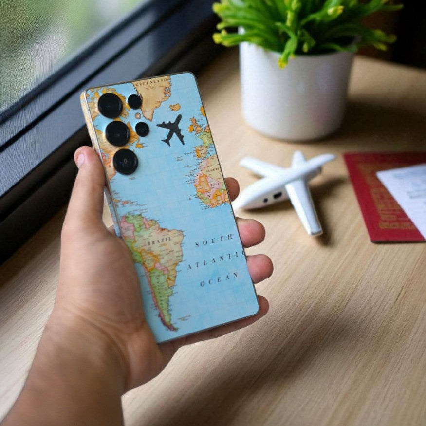 Hand holding a phone with a world map skin, airplane model, and plant in the background.